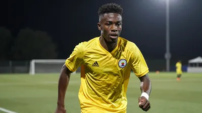 A young, athletic Black striker in his early 20s, wearing a solid yellow kit, mid-stride on a football pitch