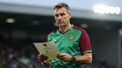 Middle-aged male club executive, athletic build, waist-up, wearing Norwich City home shirt (green body, dark red sleeves)...