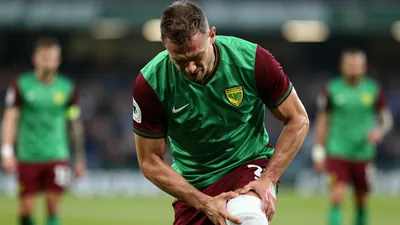 Mid-20s muscular winger, head-and-shoulders, grimacing with one hand gripping a bandaged knee, wearing Norwich City home...