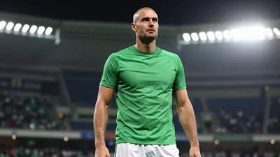 A 35-year-old, tall, and athletic-looking male coach with a strong jawline and a shaved head, wearing a bright green...
