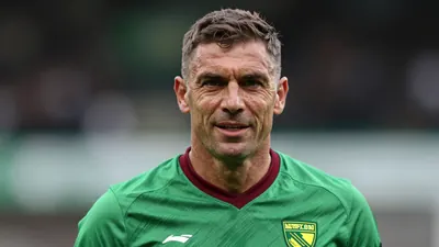 Middle-aged former defender, muscular build, mid-30s, wearing Norwich City home kit (green shirt with dark red trim...