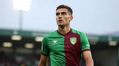 A determined, 28-year-old Norwich City midfielder with a strong build, wearing the green and dark red home kit with the...