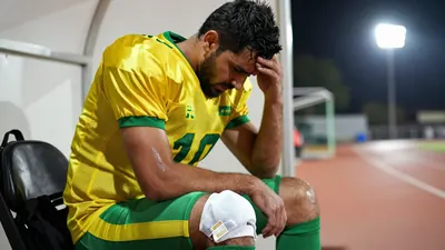 A 25-year-old male football player, wearing a yellow and green jersey, with a disappointed expression, sitting on the...