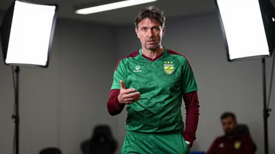 A 50-year-old, 5'11" Belgian coach, dressed in Norwich City's green and dark red home kit with the club crest on the left...