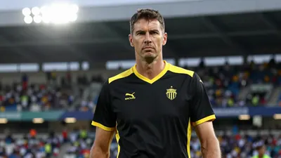 A 35-year-old, tall, and lean head coach with a serious expression, wearing a black and yellow jersey made of polyester...