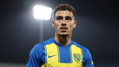 A 22-year-old winger in a Norwich City kit, standing in a training ground with a focused expression, wearing a blue and...